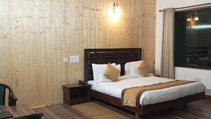 A hotel room with a wooden panel wall, a bed with white bedding and two pillows, a nightstand, a desk with a chair, and a window with curtains. - Pineland Chalets And Resort (Kasauli)