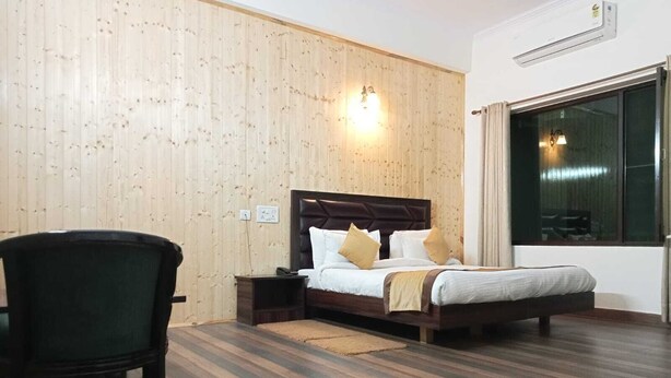 A bedroom with a wooden paneled wall, a bed with white bedding and two pillows, a nightstand, a chair, and a large window. - Pineland Chalets And Resort (Kasauli)