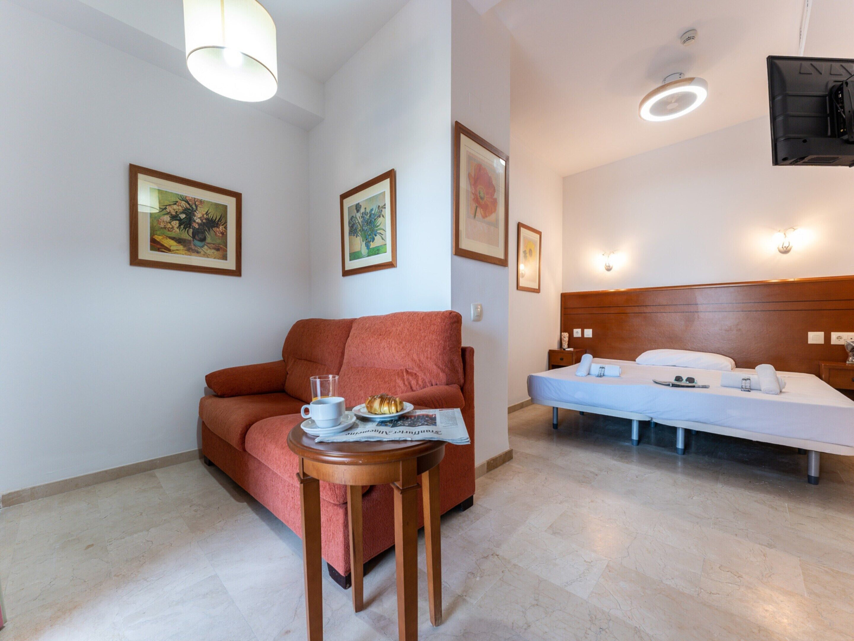 A hotel room with a bed, a sofa, a table with a cup and a pastry, and framed artwork on the walls.