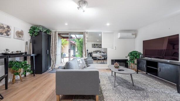 En moderne stue med en grå sofa, et sofabord og et fjernsyn på et skab. Der er et spiseområde med et sort bord og stole, samt et køkkenområde med et sort skab og en plante.