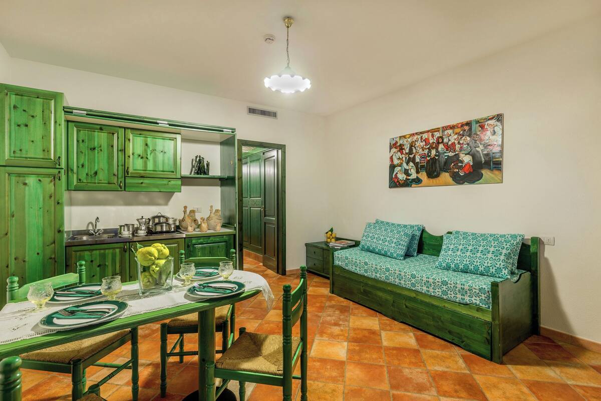 A room with a dining table set for four, a sofa, and a kitchen area with green cabinets and a hanging painting.