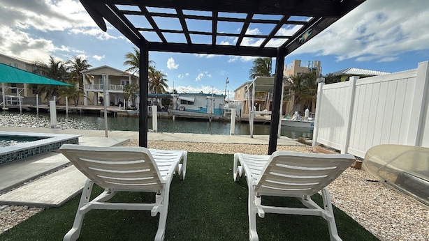 A covered outdoor area with two white lounge chairs, a pool, and a view of a marina with boats.