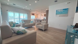 A spacious living area with a kitchen in the background, a dining table with chairs, and a large window with curtains.