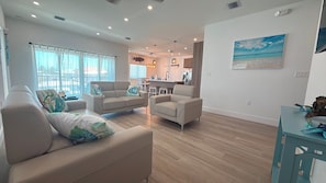 A spacious living area with a kitchen in the background, a dining table with chairs, and a large window with curtains.
