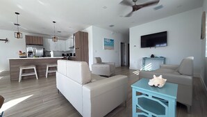 A modern living room with a sofa, a coffee table, and a TV. There is a kitchen area with cabinets and a refrigerator.