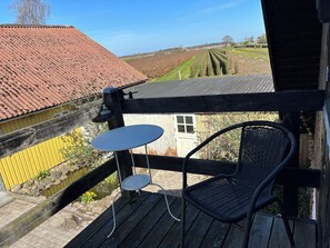 A wooden deck with a chair and a small table, overlooking a field and a building with a red roof. - Aunt Janni's Summer Pension (Bogense)