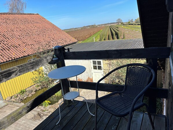 Una terraza de madera con una silla y una pequeña mesa, con vista a un campo y un edificio de techo rojo.