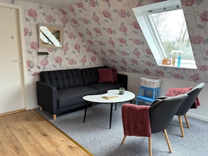 Una habitación con sofá negro, una mesita redonda, dos sillas, un ventanal con vistas y un papel tapiz floral.