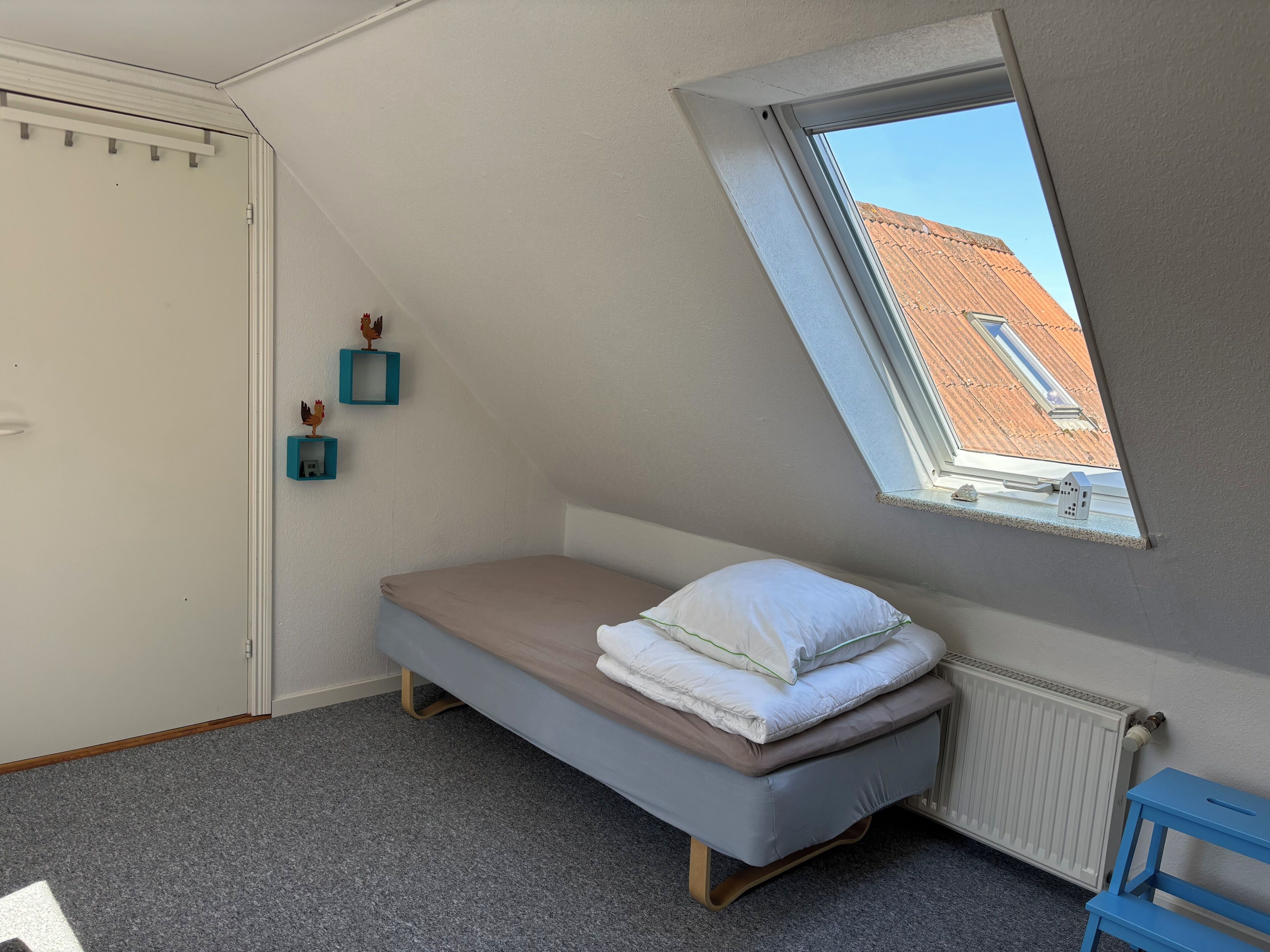 Una stanza piccola con un letto singolo, un soffitto spiovente, una finestra che si affaccia su un edificio con il tetto rosso, un termosifone e una sedia blu.