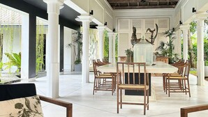Un vaste espace dédié à la salle à manger, avec une grande table entourée de chaises en bois, de colonnes blanches et de plantes en pot.