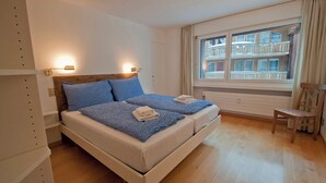 A double bed with blue bedding, a wooden headboard, and two bedside lamps. There is a window with a view of a building and a chair to the right. - Apartment Lora (Zermatt)
