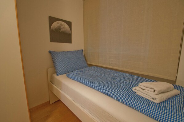 A small, single-bed room with a checkered blue and white bedspread, a white pillow, and a folded white towel on the bed. A window with a blind is on the right side, and a framed picture of a moon is on the wall to the left. - Apartment Lora (Zermatt)