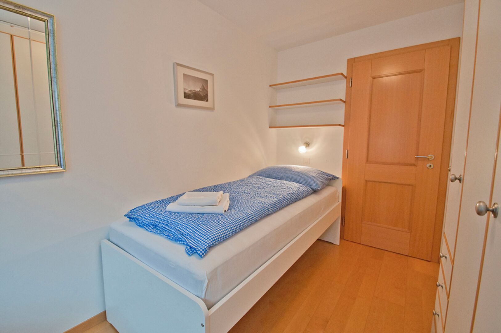 Een kleine kamer met een enkel bed, een houten deur, een nachtkastje, een wandplank en een lijst met een afbeelding.