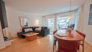 A modern living room with a sofa, coffee table, and dining table. There is a large window with curtains, a mirror on the wall, and a lamp. - Apartment Lora (Zermatt)