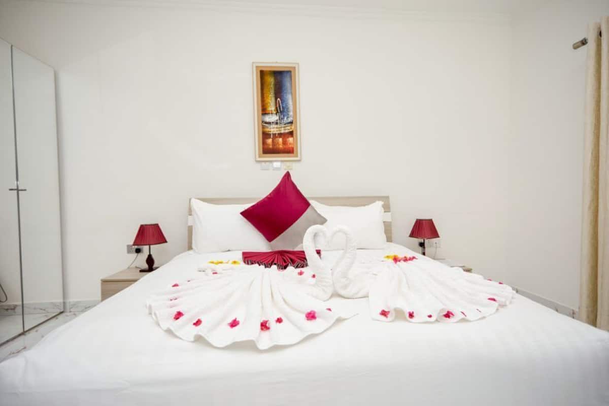 A hotel room with a large bed, two red lamps, a single red pillow, and a framed picture on the wall.