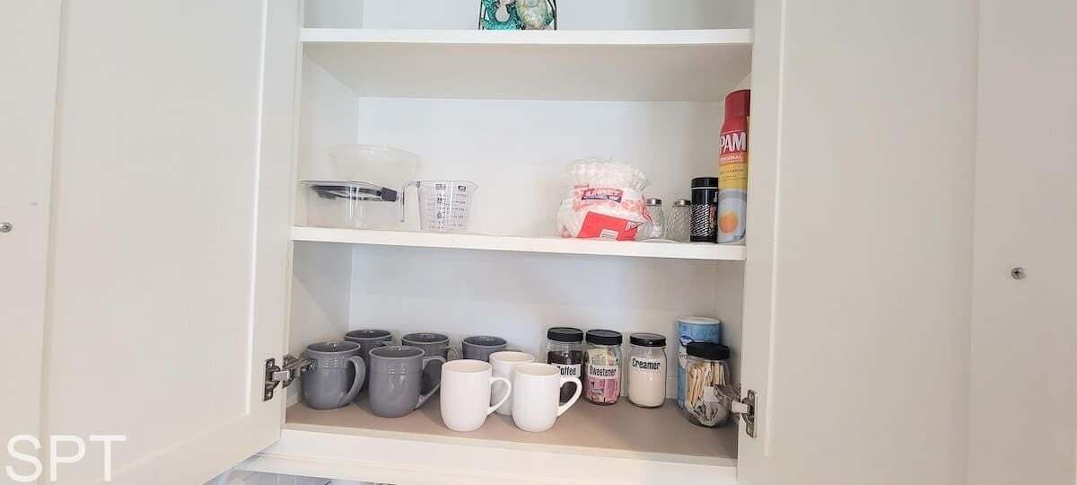 A kitchen cabinet with shelves containing various kitchen items such as coffee mugs, containers, and spices.