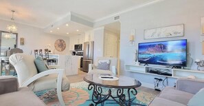 A modern living room with a TV, seating, and a kitchen in the background. - Coronado 302 2 Bedroom Home (South Padre Island)