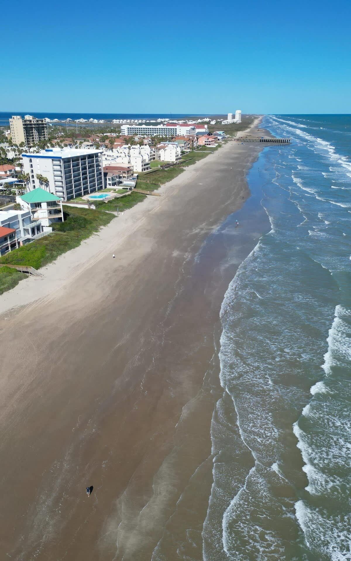 An aerial view of a beach with a long stretch of sand, a pier extending into the ocean, and a row of buildings with balconies along the shore.