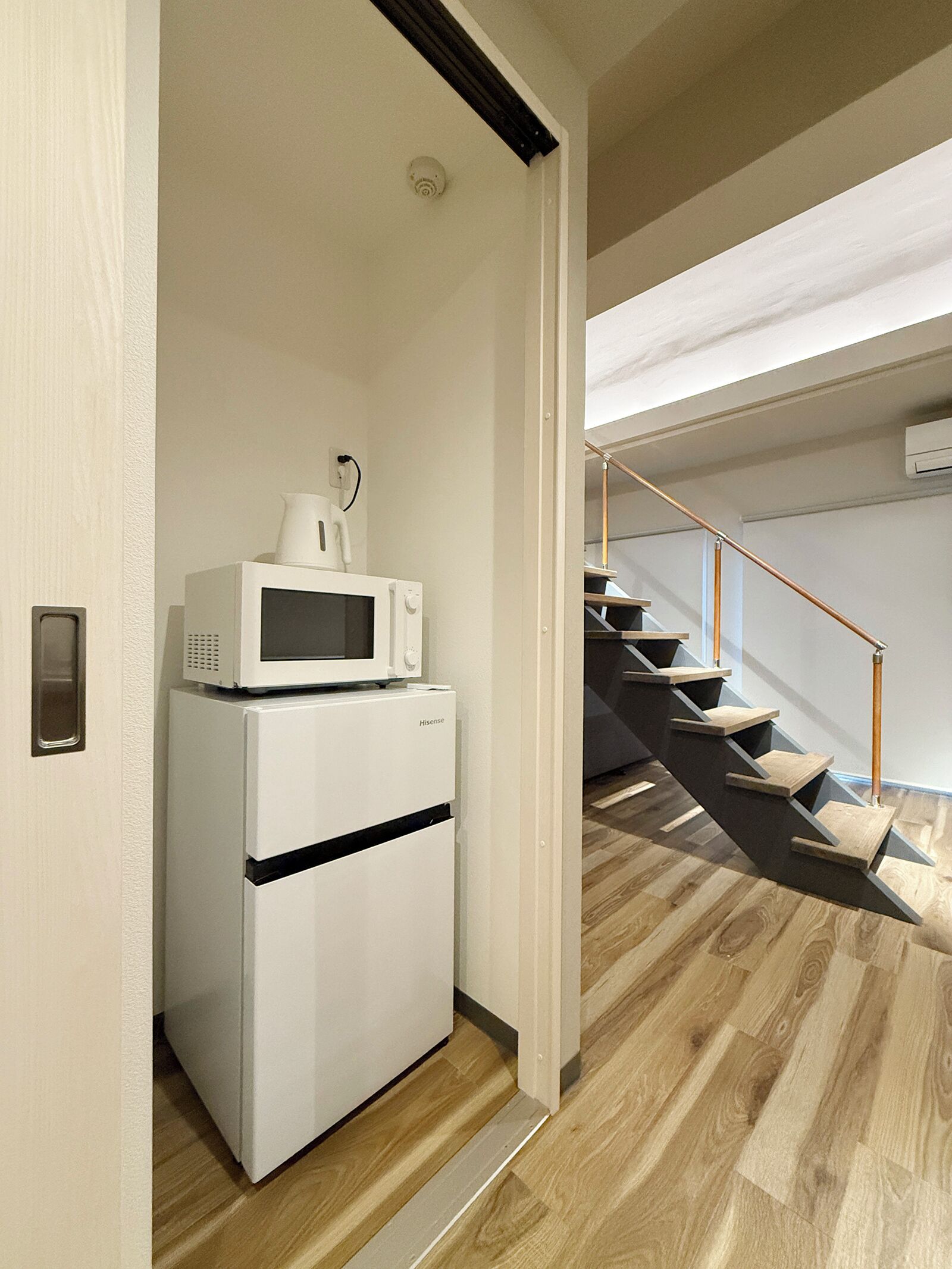 Cocina compacta con refrigerador y microondas blancos, una escalera de madera y un equipo de aire acondicionado visible.