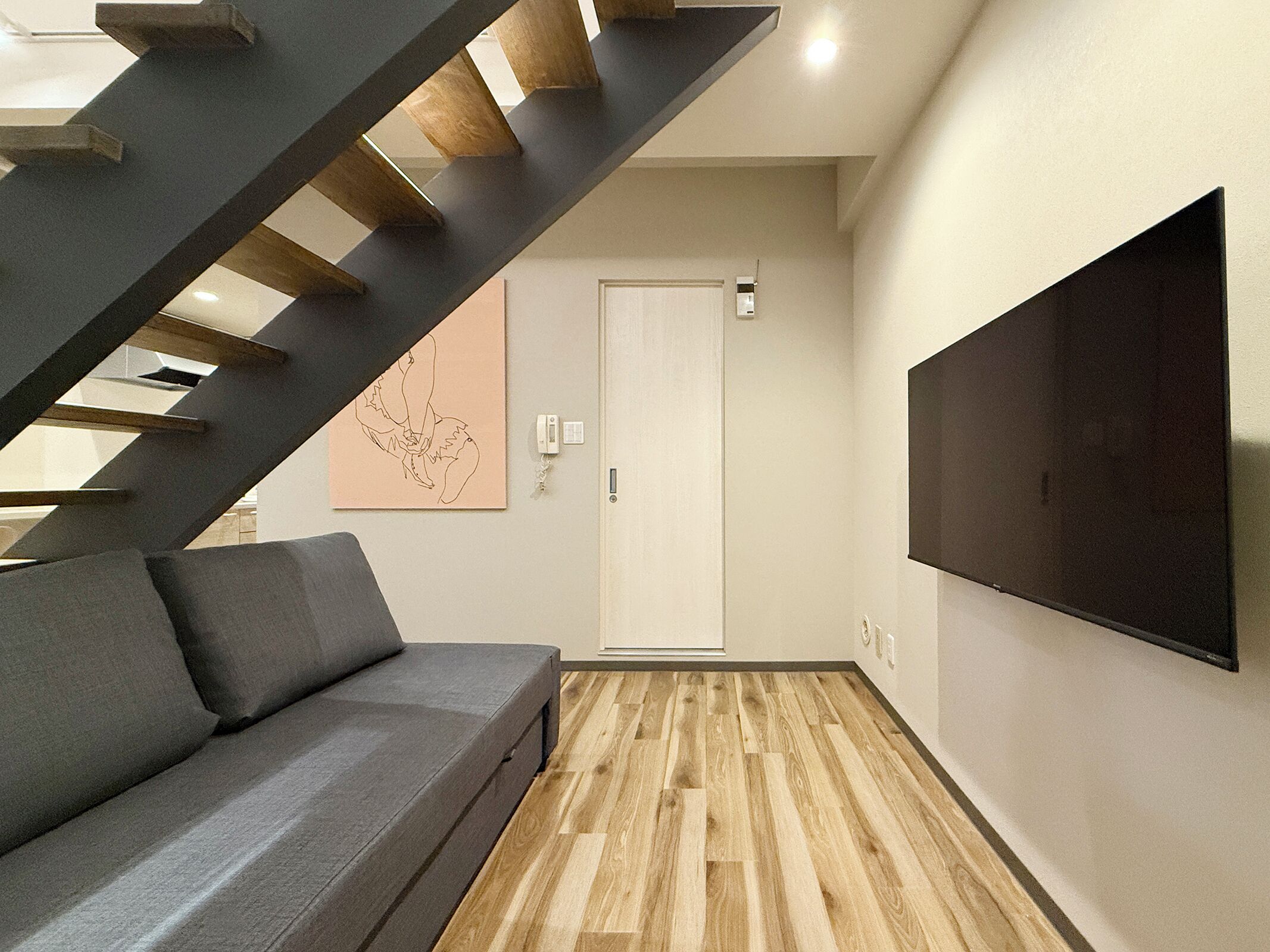 A modern living room with a grey sofa, wooden flooring, a staircase, a white door, and a mounted TV.