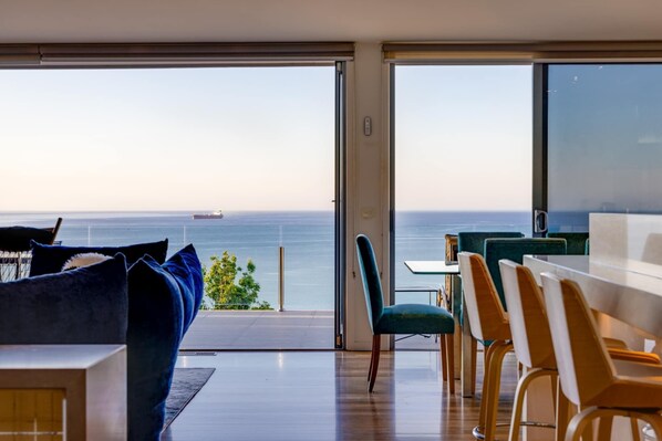 A modern interior with a large sliding glass door leading to a balcony with a view of the sea. Inside, there is a seating area with a table and chairs.