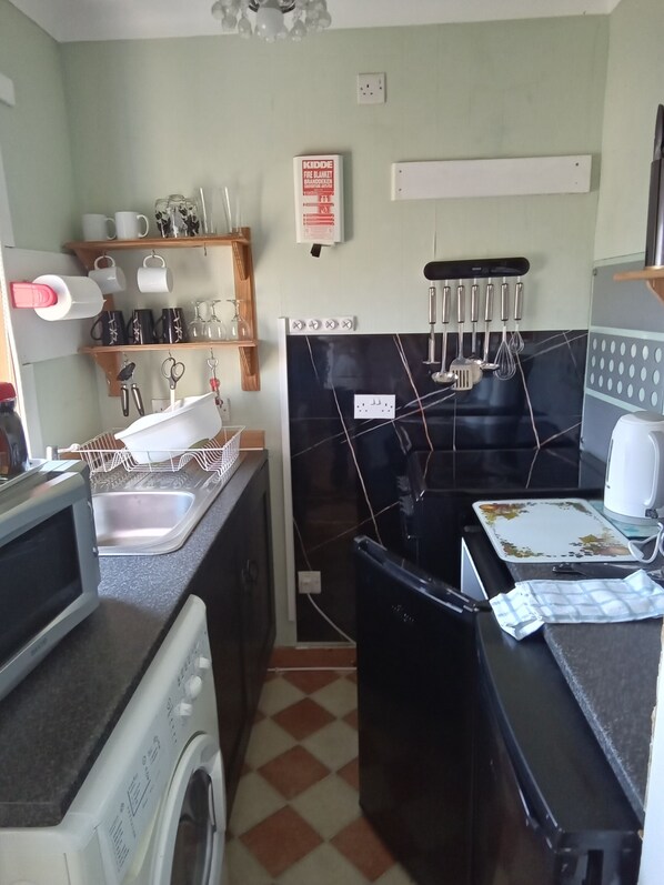 A compact kitchen with a washing machine, a microwave, and a sink. There are shelves with cups and mugs, and a fire blanket on the wall. - Small Detached Cottage in Lamlash.Sleeps 2 (Lamlash)