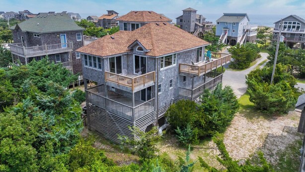 Une maison à plusieurs étages dotée d’un balcon et d’une terrasse, entourée d’arbres et bordée d’une zone sablonneuse.