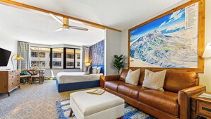 A hotel room with a bed, a brown leather sofa, a white ottoman, a desk with a lamp, a television, and a large map on the wall. - Grand Lodge 412 & 414 — Pets OK | Hot Tub | Pool | Sleeps 8 (Crested Butte)