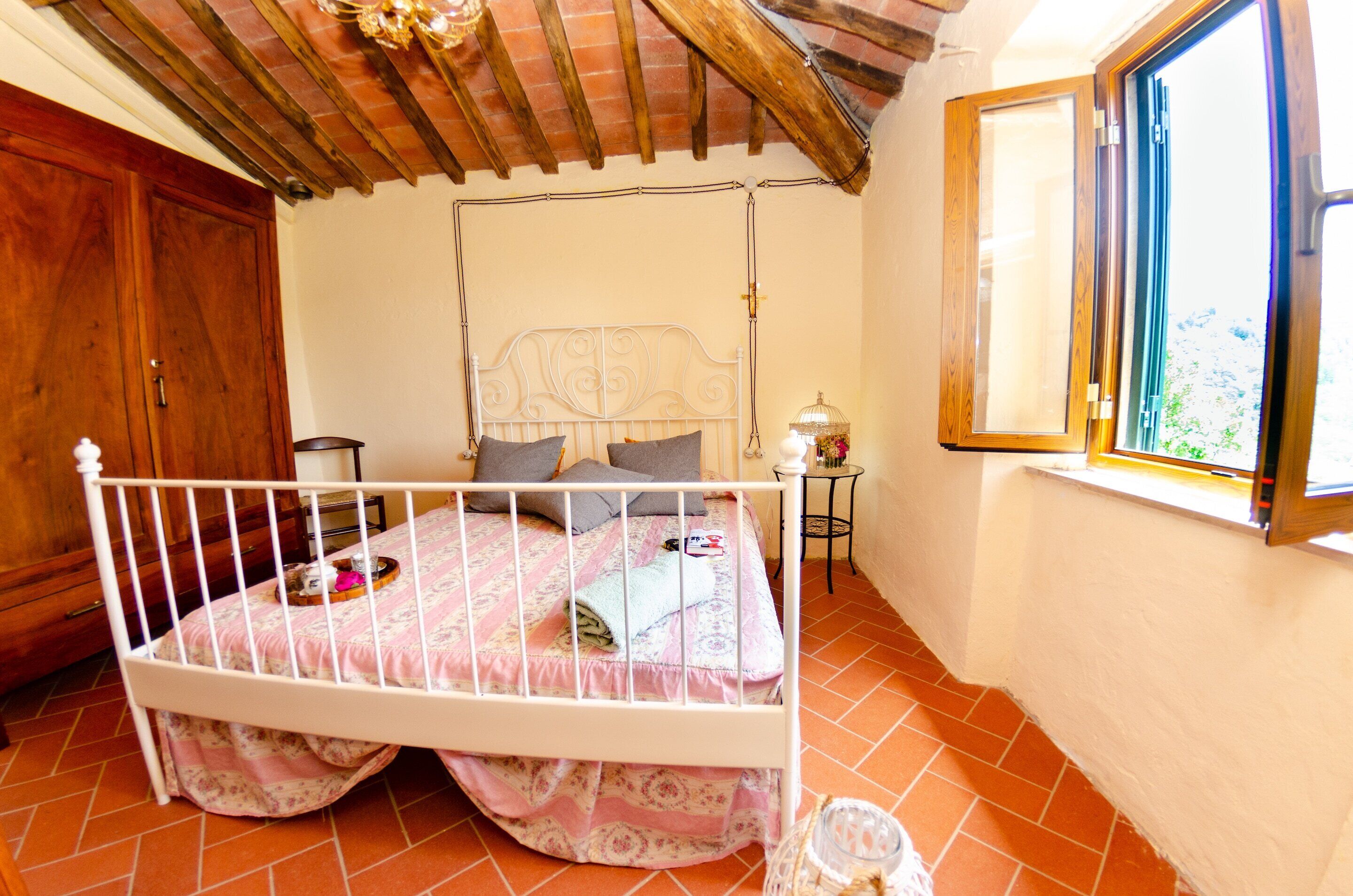 A bedroom with a white metal bed frame, a wooden wardrobe, a window with a view, and a tiled floor.
