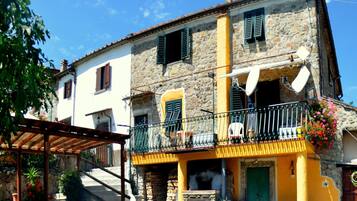 A two-story stone house with a yellow facade, a balcony with a metal railing, and a small outdoor seating area.