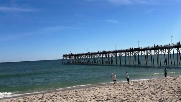 Una playa con un muelle que se adentra en el agua, arena y cielo despejado.