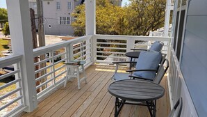 Una terraza de madera con baranda blanca, una mesa con dos sillas y vistas a casas vecinas.