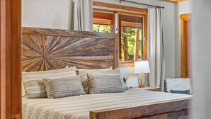 A bed with a wooden headboard, a bedside table with a lamp, and a view of trees through a window.
