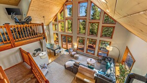 A spacious loft-style living area with a large window overlooking a forest, a dining area, and a staircase leading to an upper level.