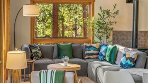 A cozy living room with a sectional sofa, a round wooden coffee table, a lamp, and a large window with a view of trees.