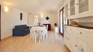 A dining area with a white table and chairs, a blue sofa, and a kitchen with white cabinets and a fruit bowl. - Villas with common or private pool in Costa Rei (Muravera)