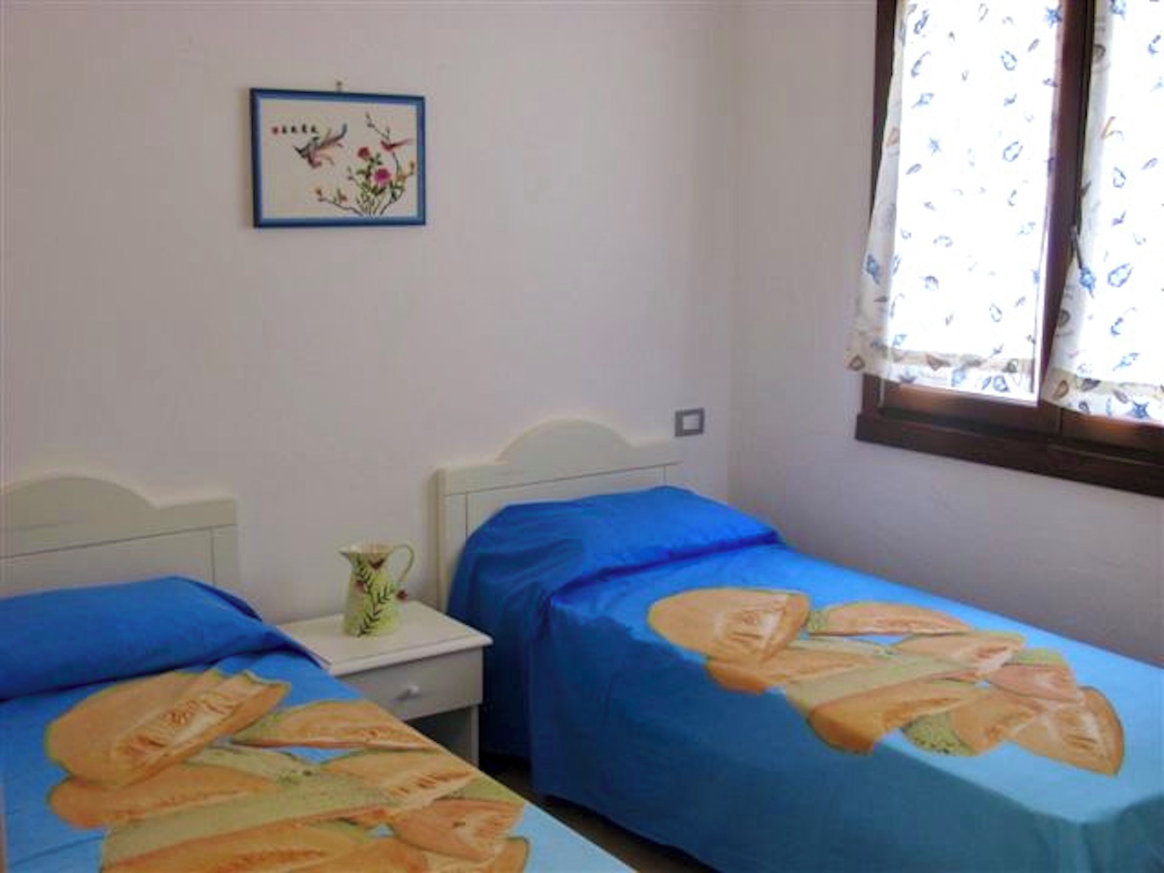 A bedroom with two beds, a nightstand, a framed picture on the wall, and a window with patterned curtains.