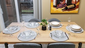 Een eettafel gedekt met borden, glazen en bestek, met een schilderij van wijnflessen aan de muur.