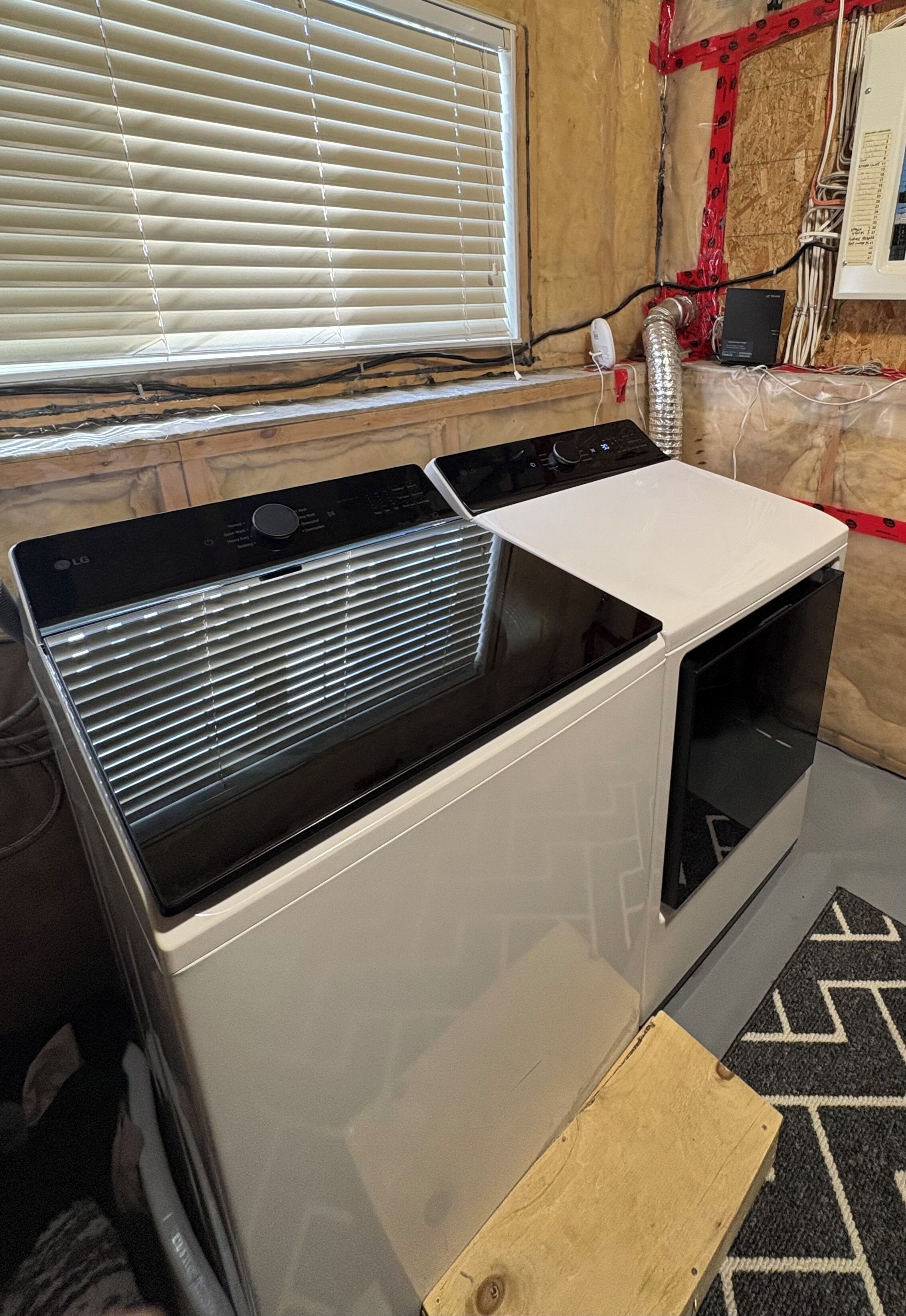Un appareil compact combinant lave-linge et sèche-linge, doté d’un panneau de commande, installé dans une pièce dont les murs sont recouverts de lambris et qui possède une fenêtre avec des stores.