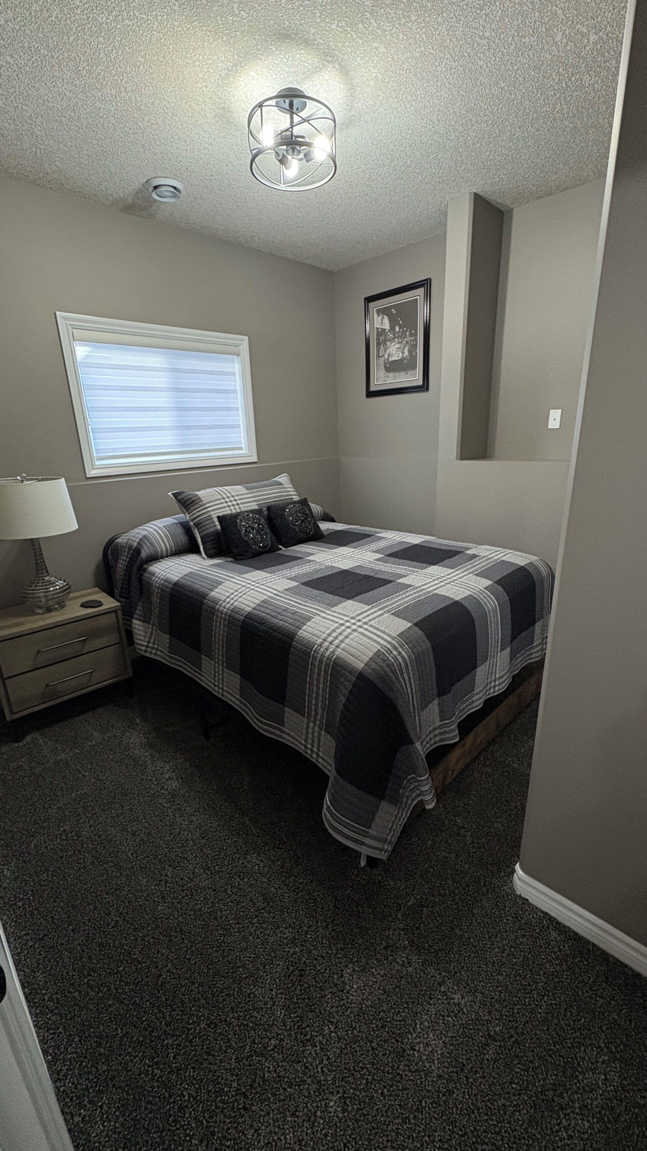 Une chambre à coucher comprenant un lit, une table de chevet, une lampe, une fenêtre avec des stores, un luminaire au plafond et un tableau encadré accroché au mur.