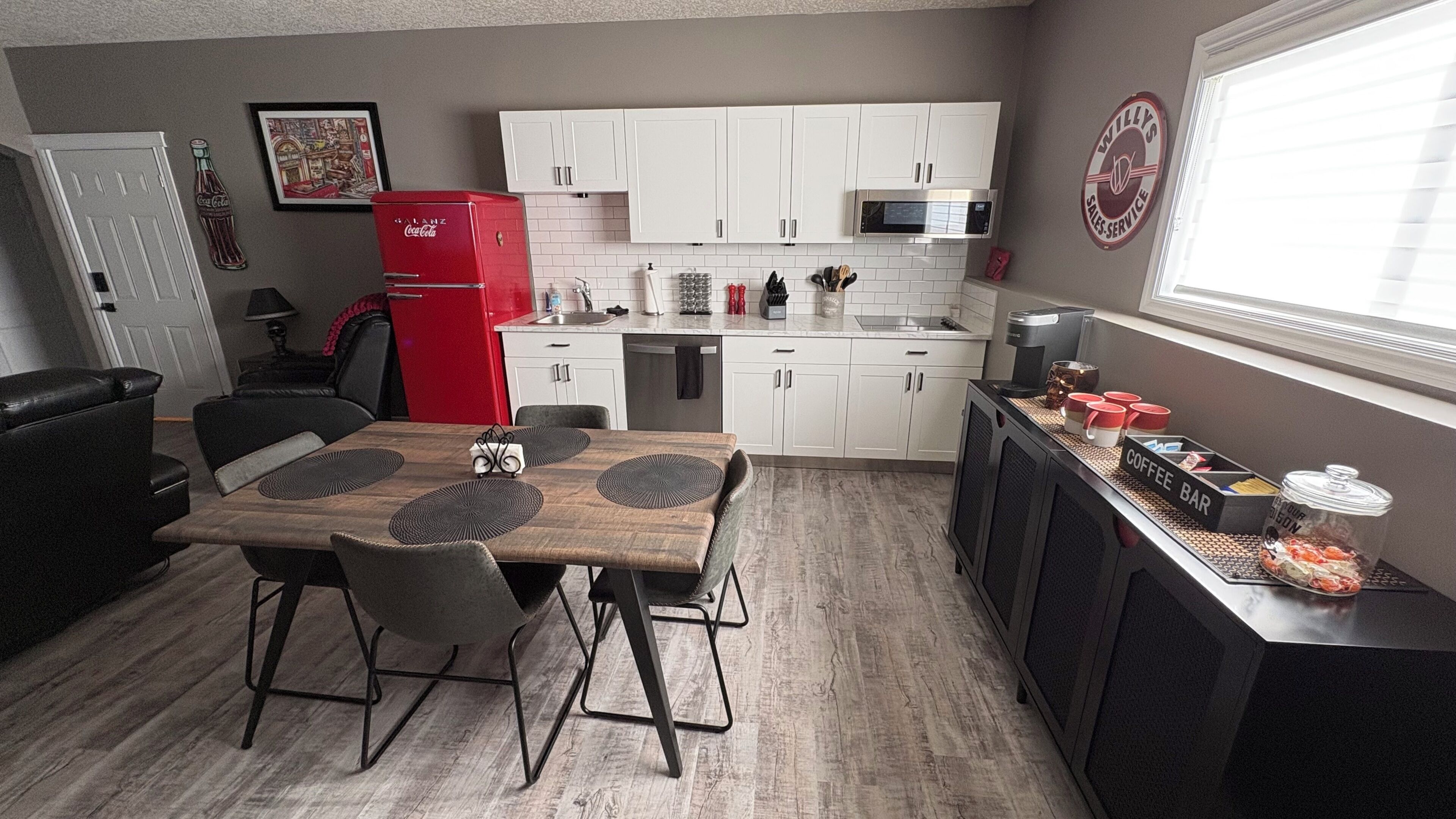 A compact kitchen with white cabinets, a red refrigerator, a coffee bar, and a dining area with a wooden table and chairs.