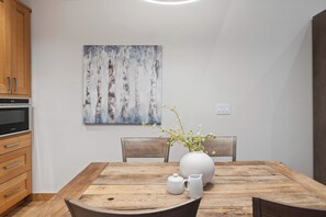 Un comedor con una mesa de madera, sillas, un jarrón blanco con flores, una jarra blanca y un cuadro en la pared.
