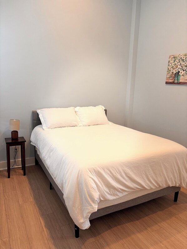 Una cama bien hecha con ropa de cama blanca en una habitación con suelo de madera y una mesita de noche pequeña con una lámpara.