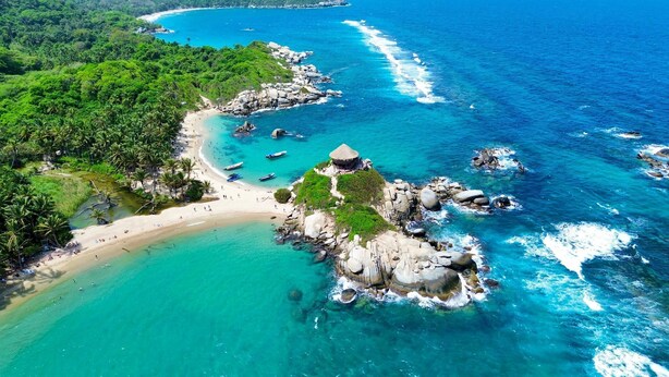 Vista aerea di una spiaggia tropicale con acque cristalline turchesi, affioramenti rocciosi e vegetazione lussureggiante.
