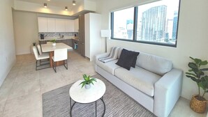 A modern apartment with a kitchen, dining area, and living room. It features a large window with a city view, a sofa, a coffee table, and a potted plant.