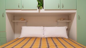 Una cama con almohadas blancas y una colcha a rayas amarilla y marrón, armarios verdes y dos pequeñas estanterías con objetos decorativos.