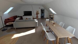 A modern living room with a sofa, dining table, and chairs. There is a TV and a bookshelf in the background. - La Canopée des Arbres Juliette! Relax and enjoy yourself at the edge of the forest! (Ellezelles)