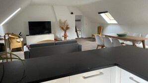 A modern kitchen with a black countertop and white cabinets, a dining area with a wooden table and chairs, and a TV mounted on the wall. - La Canopée des Arbres Juliette! Relax and enjoy yourself at the edge of the forest! (Ellezelles)