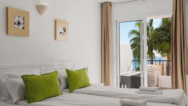 A hotel room with a bed, two green pillows, a white headboard, a wall-mounted light, and a balcony with a view of palm trees. - REF 96 - Charming Apartment with Pool Near Beaches & Golf Courses (Mijas)