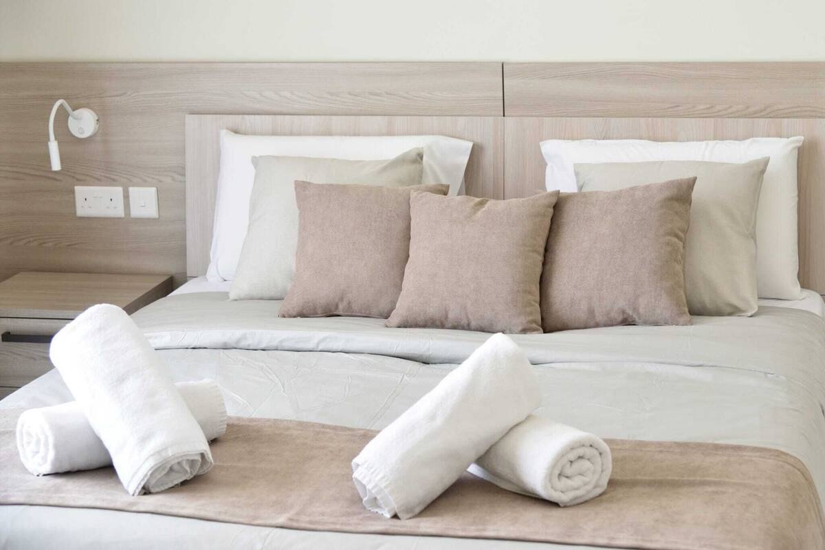 Un letto rifatto con cura, con biancheria beige e bianca, una testiera in legno e un comodino con lampada e prese elettriche.